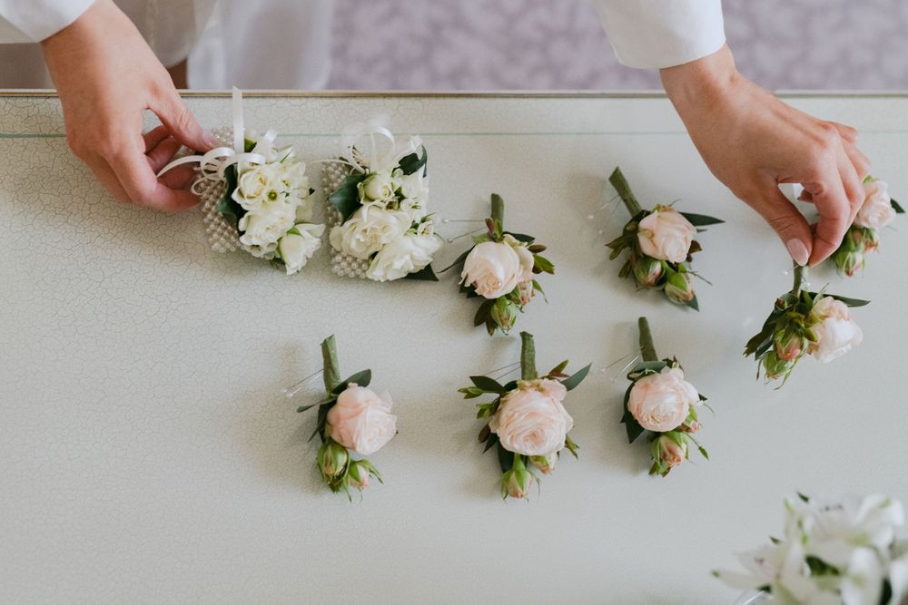 A row of delicate pink and white rose boutonnieres lined up on a light surface with hands arranging them.