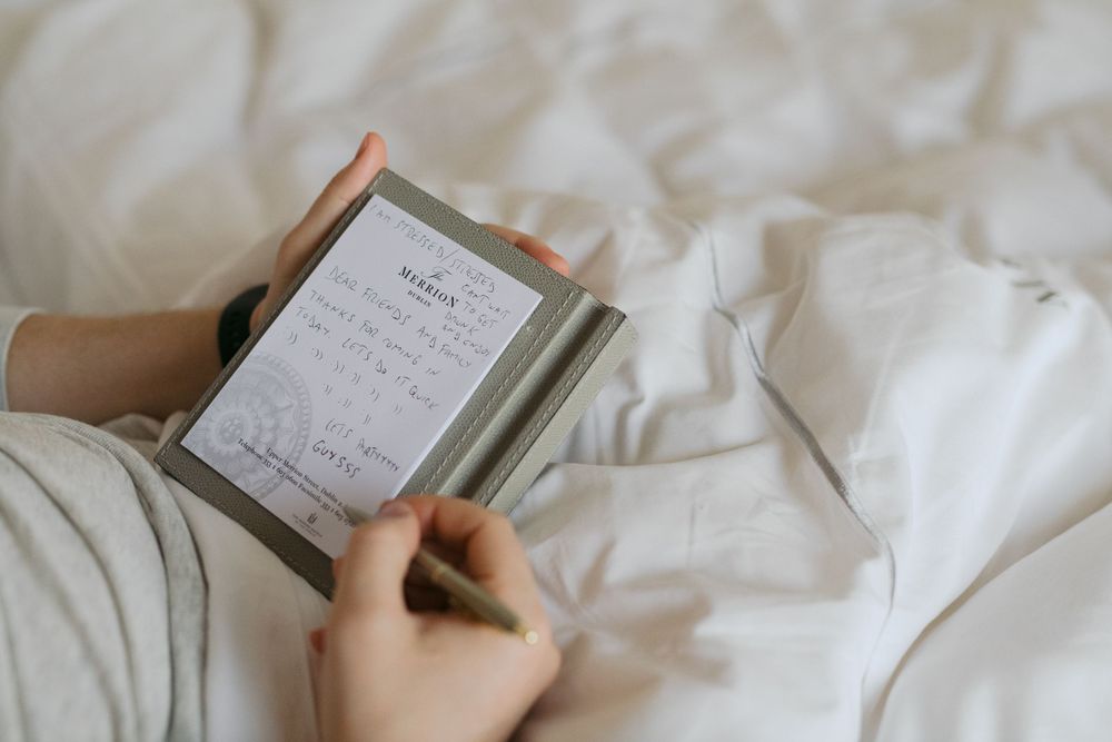 Hands holding a notebook and pen while writing on crisp white bedsheets.