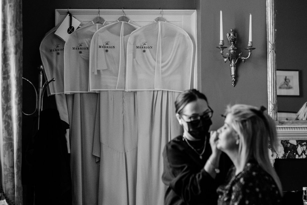 A black and white photo of a makeup application session with wedding dresses hanging in the background.