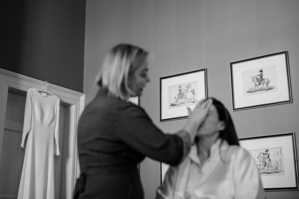 A makeup artist applies cosmetics to someone sitting in a chair while art prints hang on the gray wall behind them.