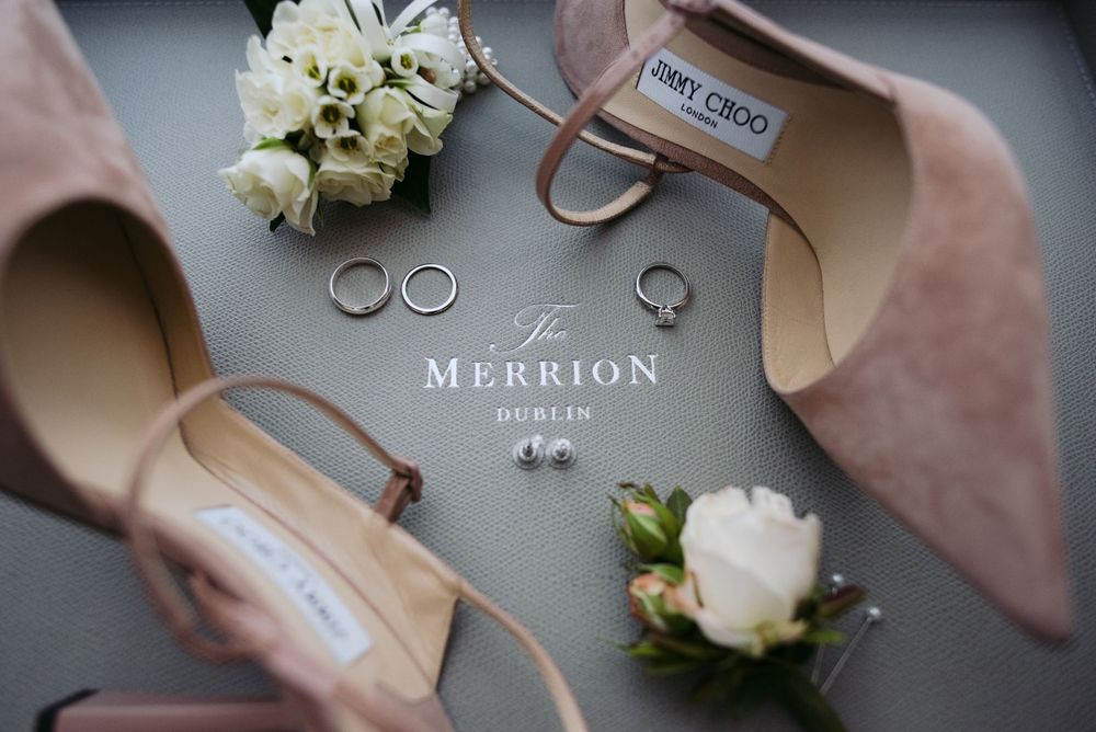 Elegant wedding rings and bouquet arranged with Jimmy Choo shoes and Merrion branding on gray background.