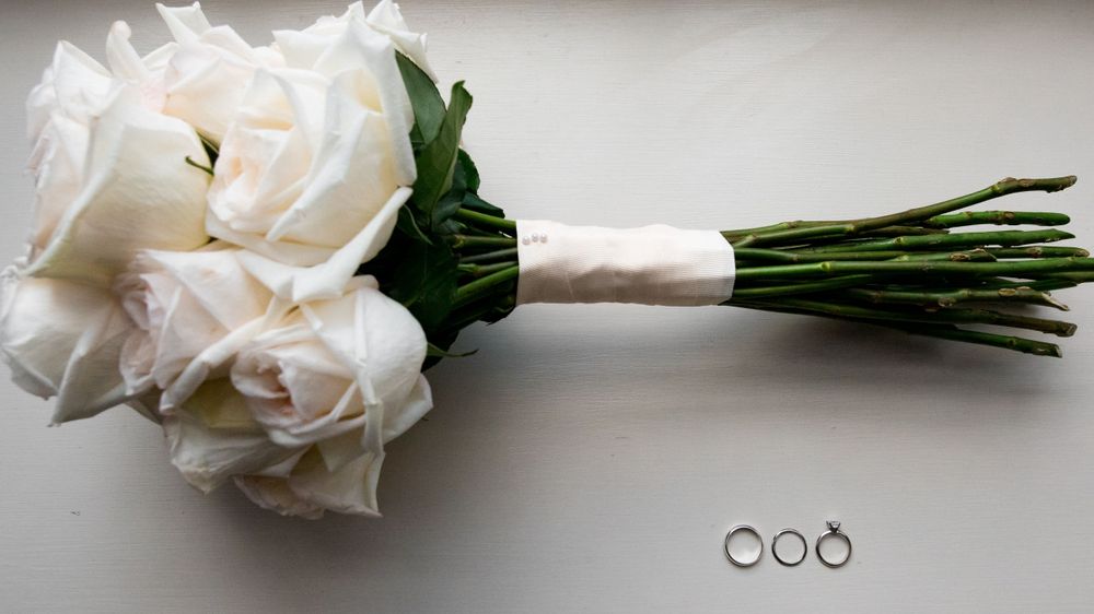 Classic white rose wedding bouquet with three silver rings arranged against a light gray background.