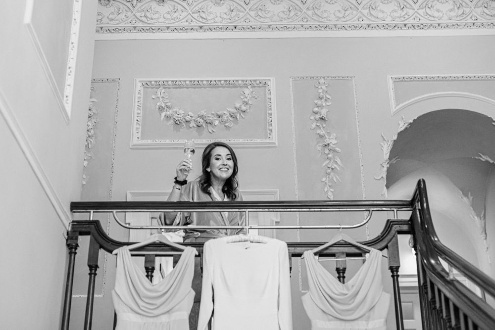 A joyful bride stands at an ornate balcony railing in a historic venue, captured in elegant black and white photography.