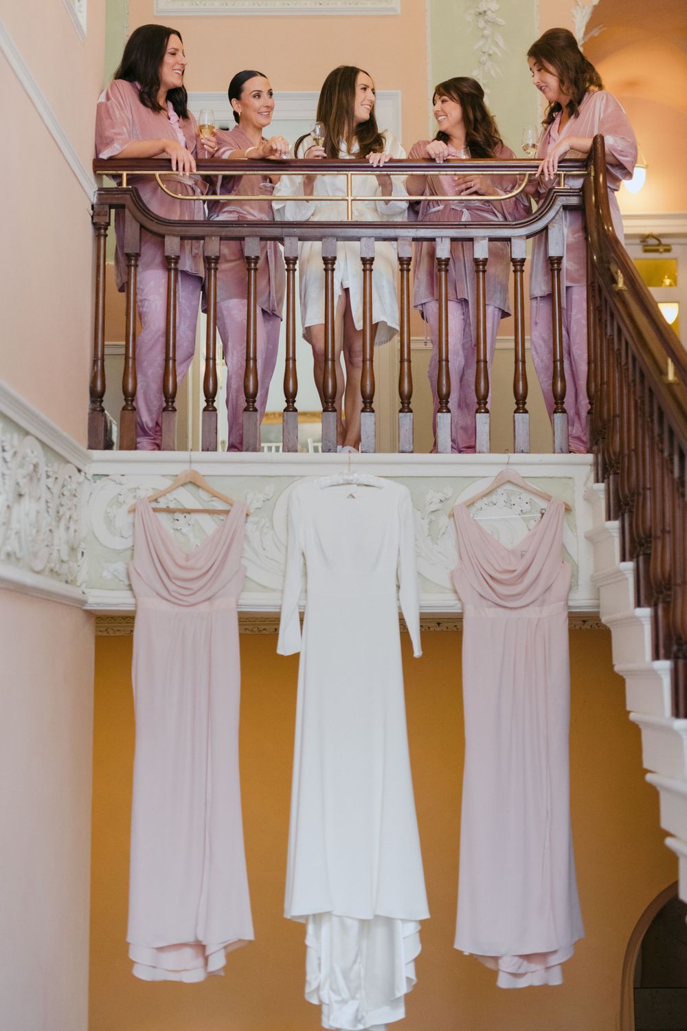 Wedding party gathers on staircase with bridal dresses hanging below in white and blush pink.