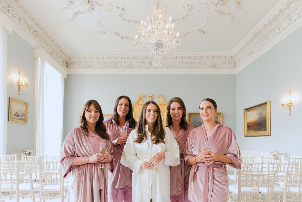 Bridal party in matching pink robes stands together in an elegant room with ornate white ceiling and wall sconces.