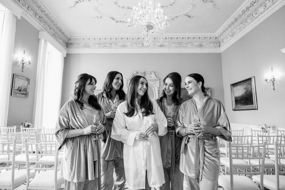 A group of people wearing matching robes laugh together in an elegant room with ornate moldings and wall sconces.
