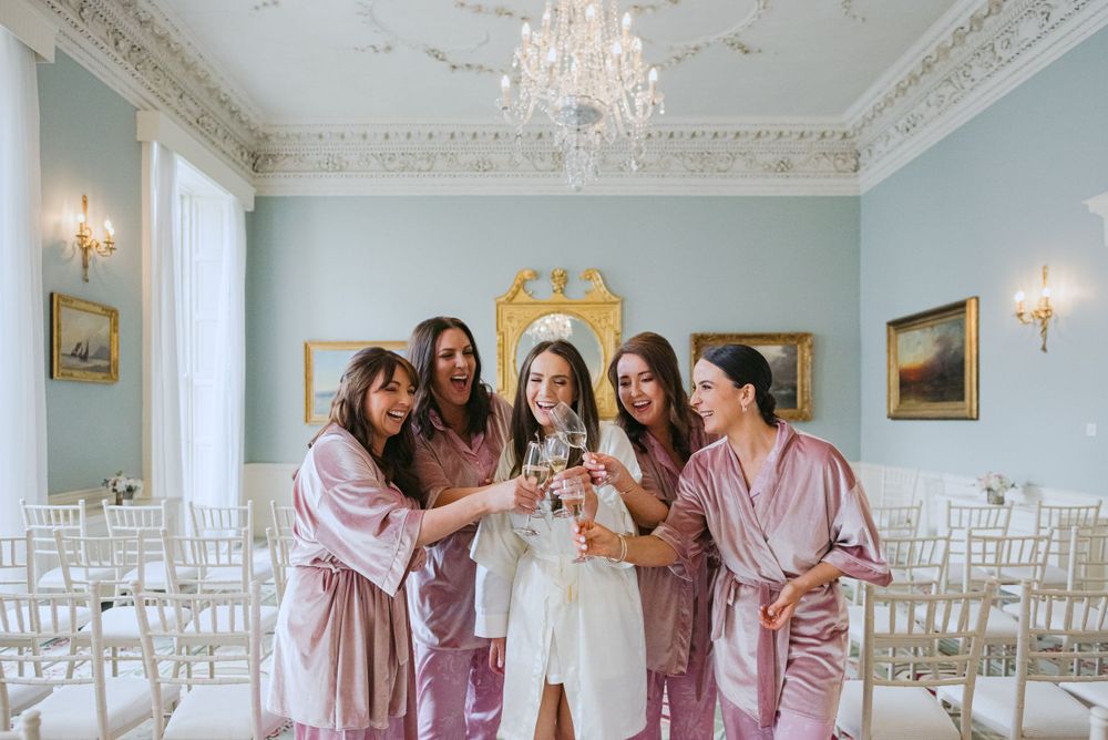A group wearing matching pink satin robes gather closely together with champagne glasses in an elegant room with chandeliers.