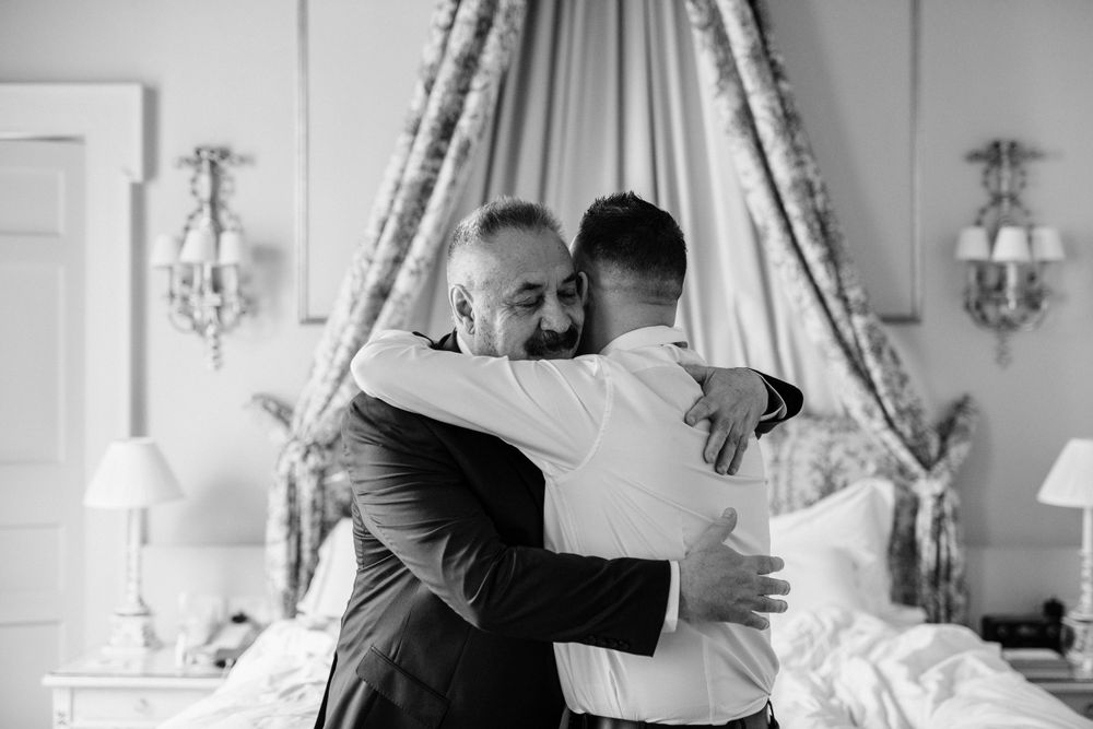 A tender emotional embrace shared between two people in a bright hotel room with elegant curtains and wall sconces.