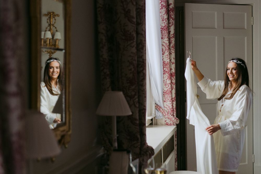 Ethereal morning light streams through curtains as someone in a white robe gets ready in a bedroom with ornate decor.
