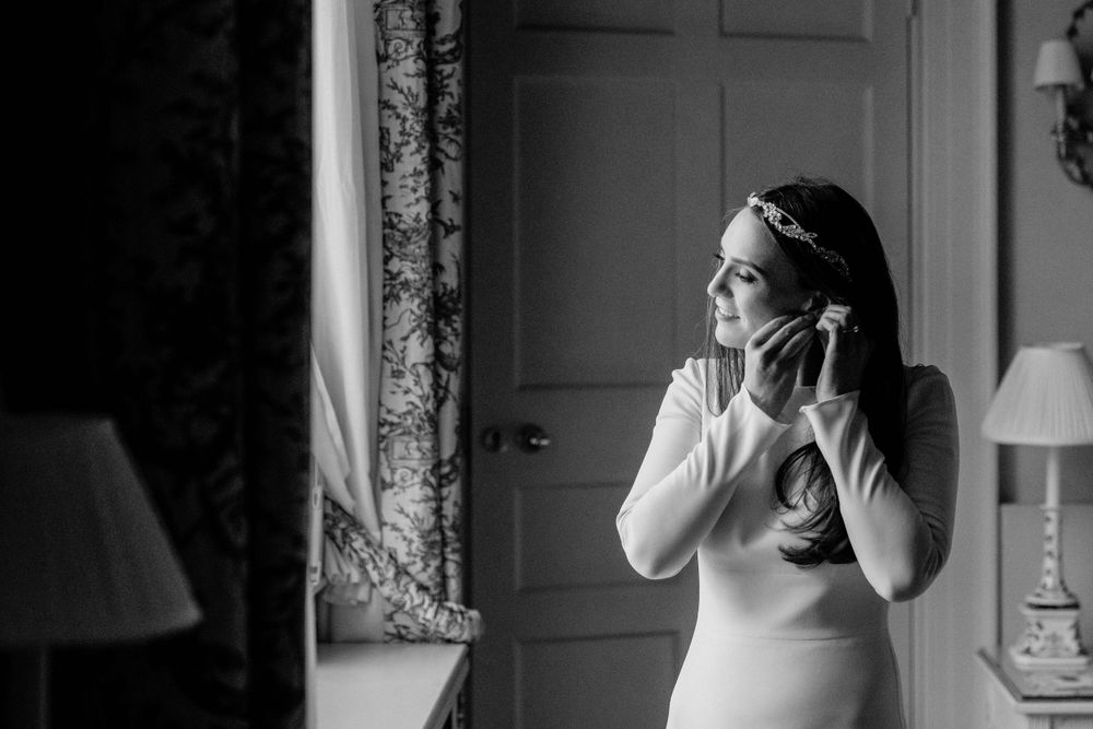 Black and white image of someone in a formal white dress standing near a window and holding a phone.