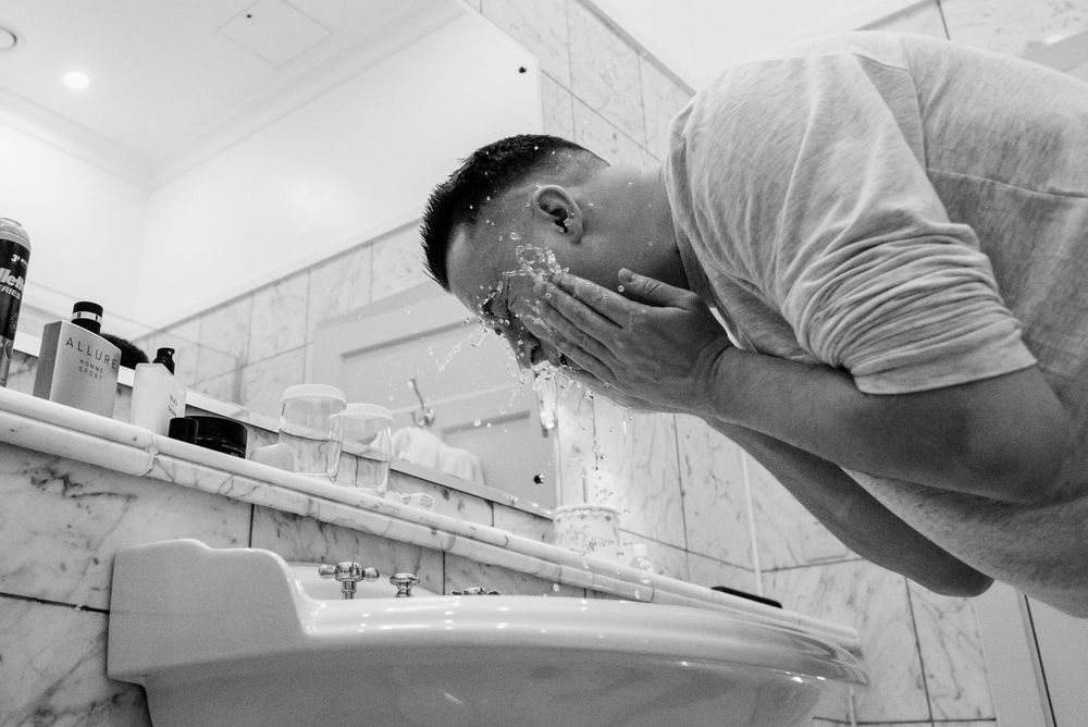 A person leaning over a porcelain sink with hands over their face in a monochrome image.