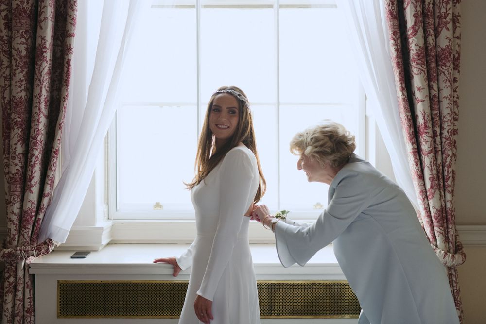 Two people stand by a bright window with flowing curtains during a dress fitting moment.