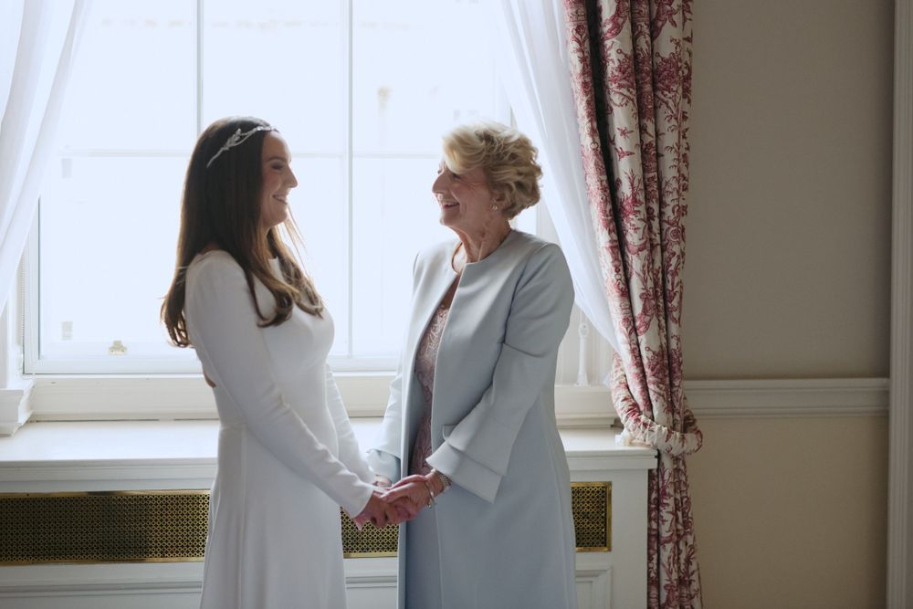 Two women stand together by a window holding hands, one wearing white and one in a light gray blazer.