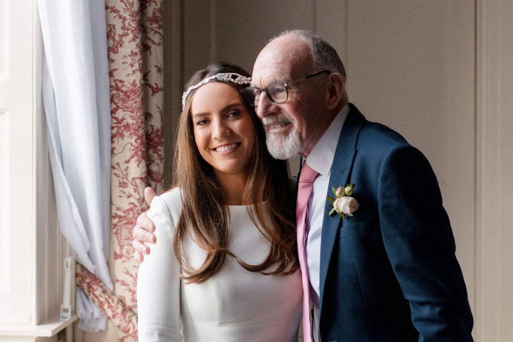 Two attendees at a wedding event stand together by a window, one wearing a white dress with floral crown and one in navy suit.