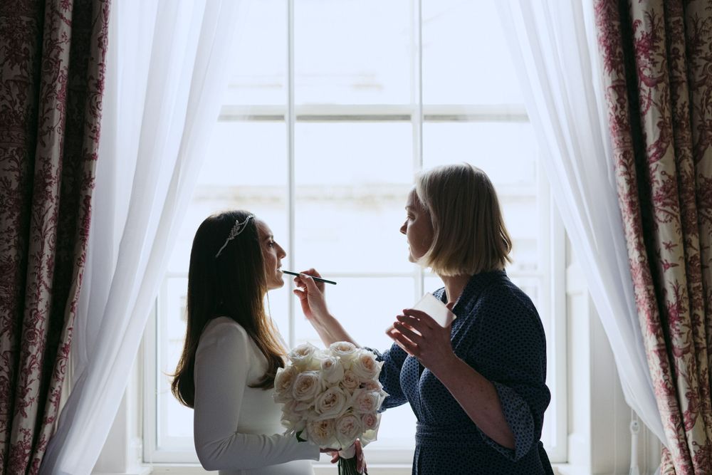 Two people in front of window with white curtains during makeup application session.