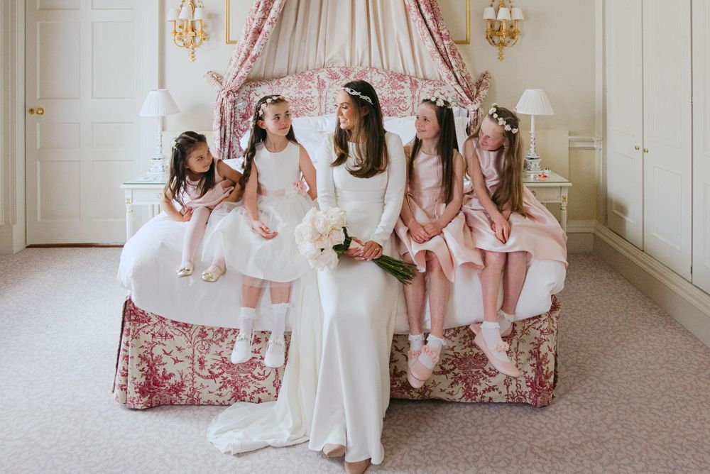 A bride and bridesmaids in white and pink dresses sit together on an elegant floral couch in a luxurious room.
