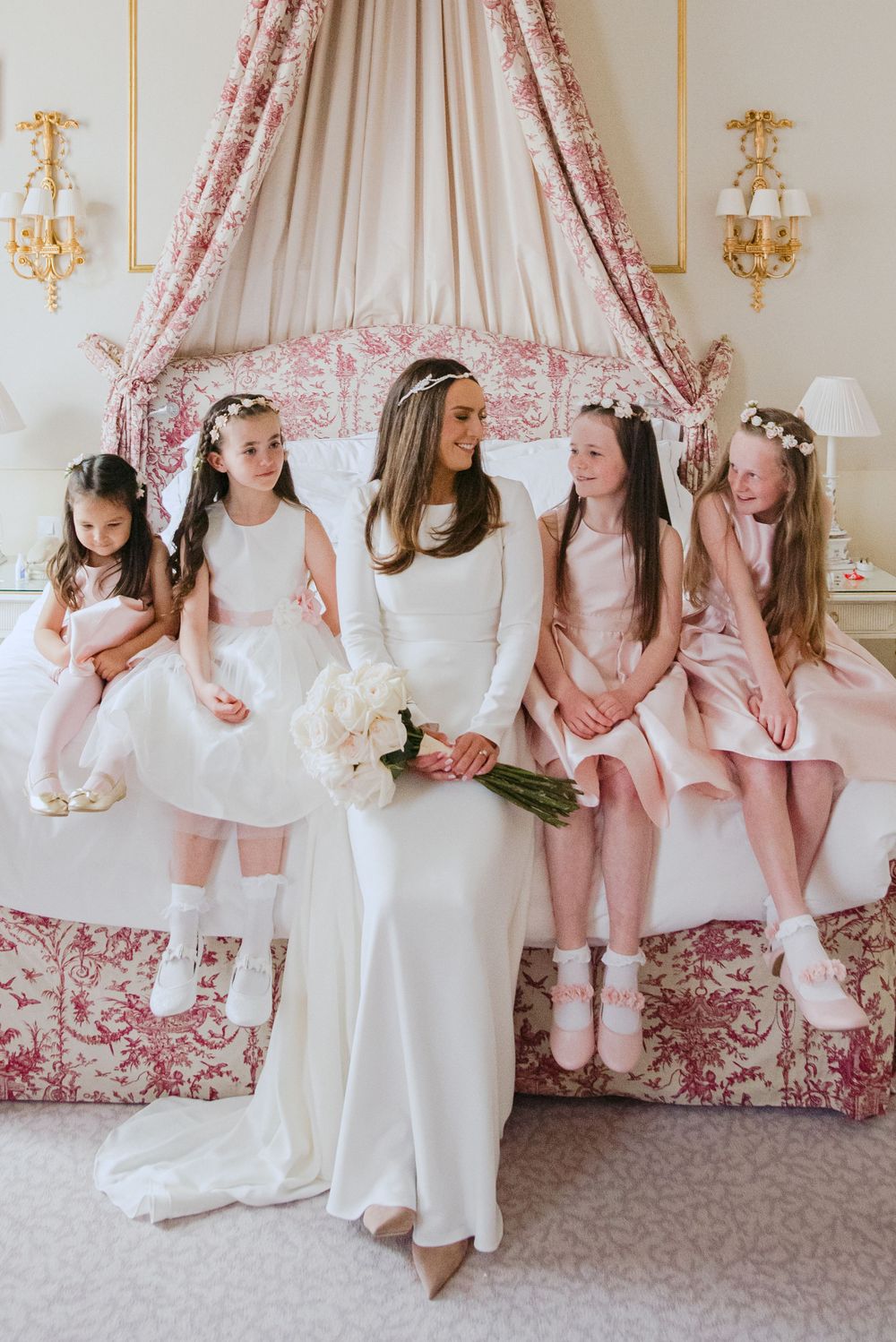 A group in white and pink dresses sit together on an elegant bed with pink toile fabric and gold wall sconces.