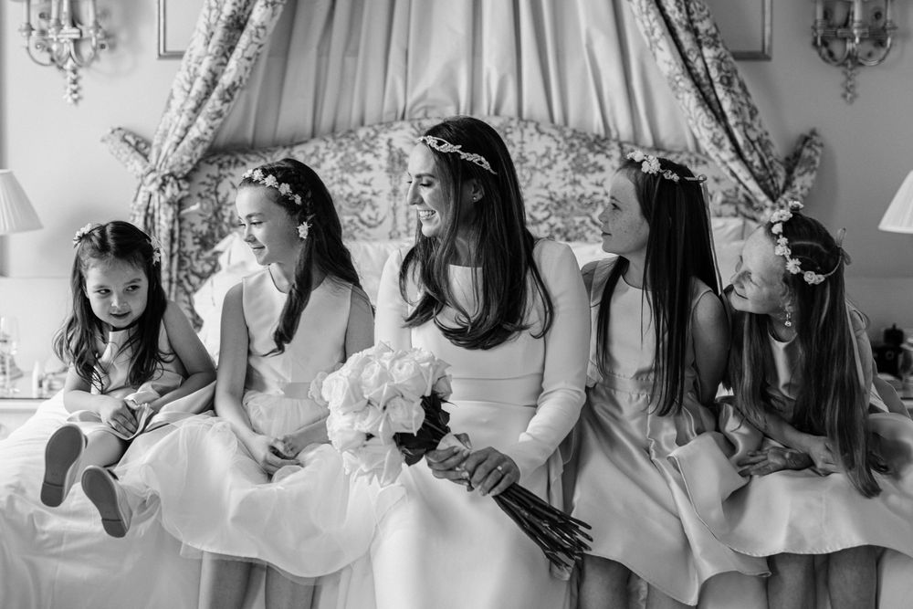 Young bridesmaids in white dresses and floral crowns sit together on an elegant vintage sofa during a wedding celebration.