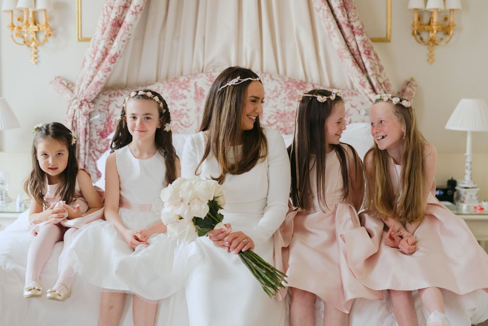A bride and flower girls in white and pink dresses sit together on a couch in an elegant room with floral curtains.