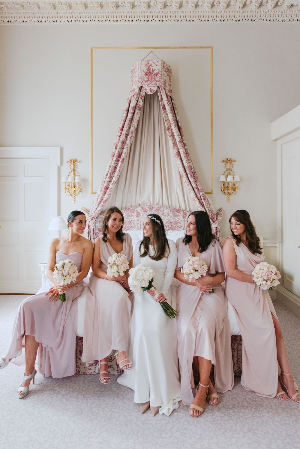 Bridesmaids in blush pink dresses sit together under an ornate canopy in an elegant white room with wall sconces.