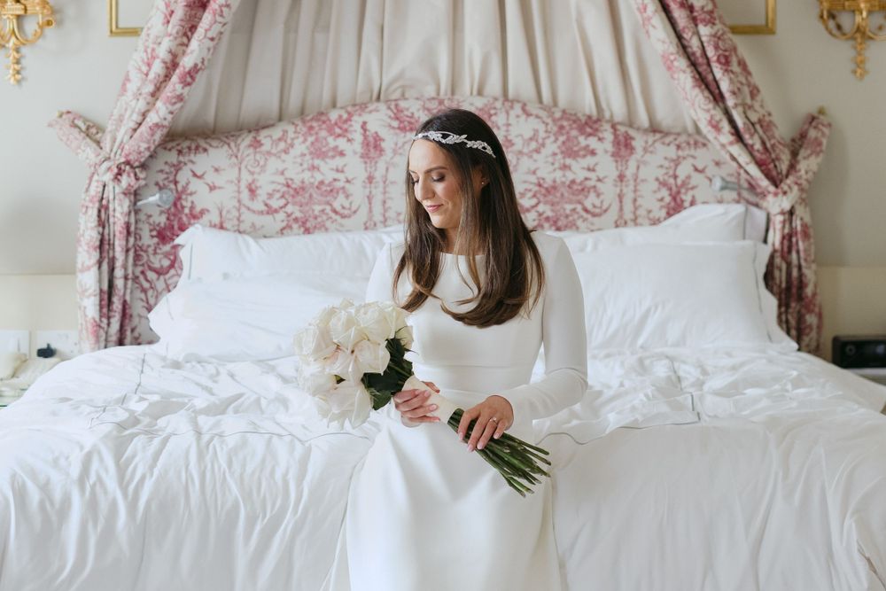 A bride in a classic white long-sleeved gown sits on a bed with white linens holding a bouquet under an ornate canopy.