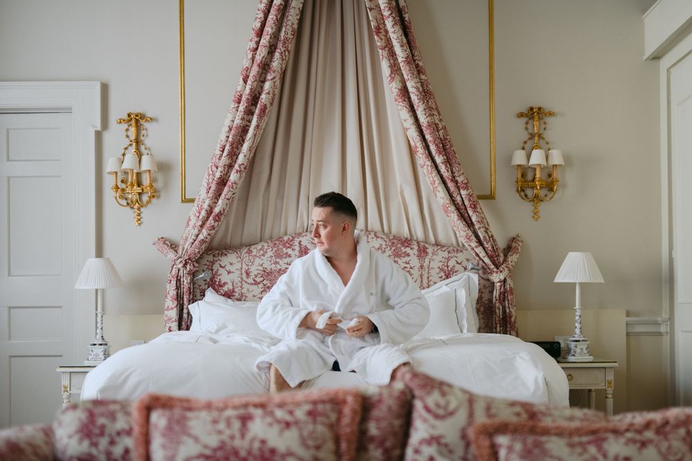 A person wearing a white bathrobe sits in an elegant bed with a floral canopy and gold wall sconces in an upscale hotel room.