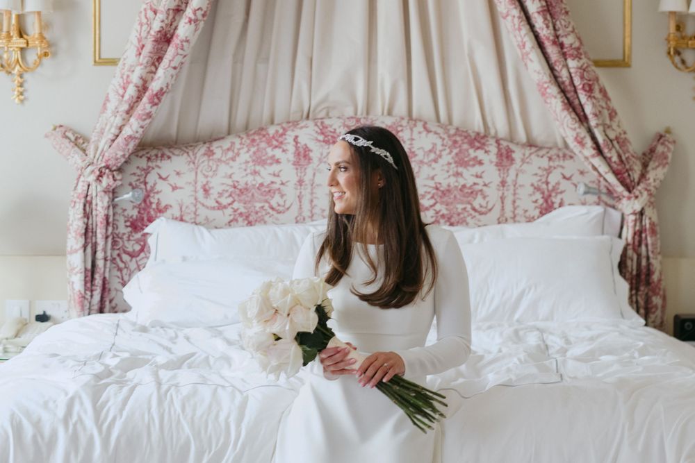 Elegant scene showing a person in white dress sitting on bed with pink floral canopy and holding white roses.