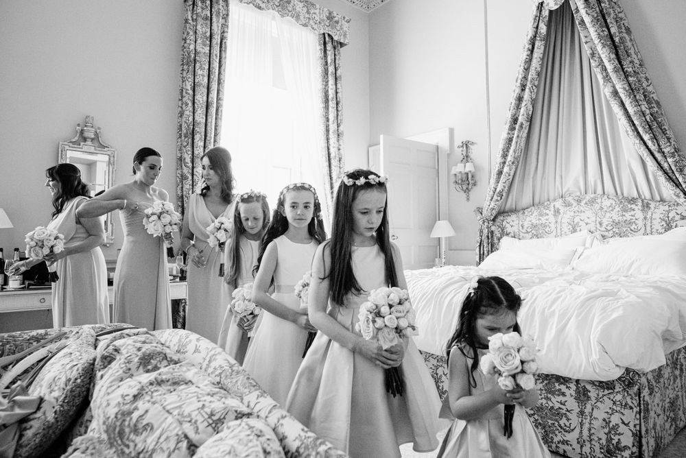 Young bridesmaids in elegant white dresses gather in an elegant room with floral bouquets during wedding preparations.