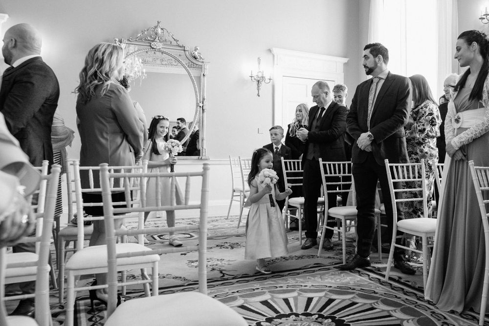 A black and white wedding ceremony photo showing a young attendant walking down the aisle past white chiavari chairs.