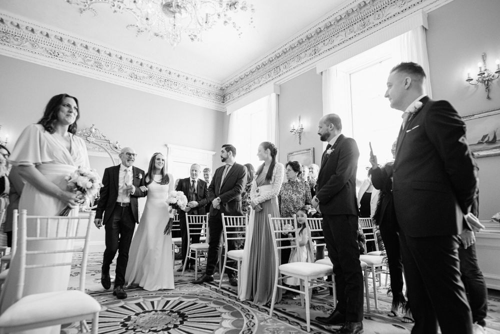 Wedding guests gather in an elegant room with ornate trim and wall sconces for a celebration in black and white.