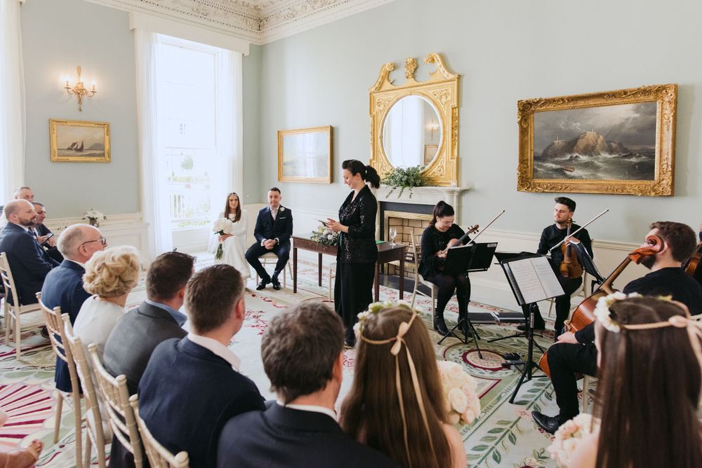 Musicians perform for an elegant audience in an ornate white room with gold-framed mirrors and artwork on the walls.