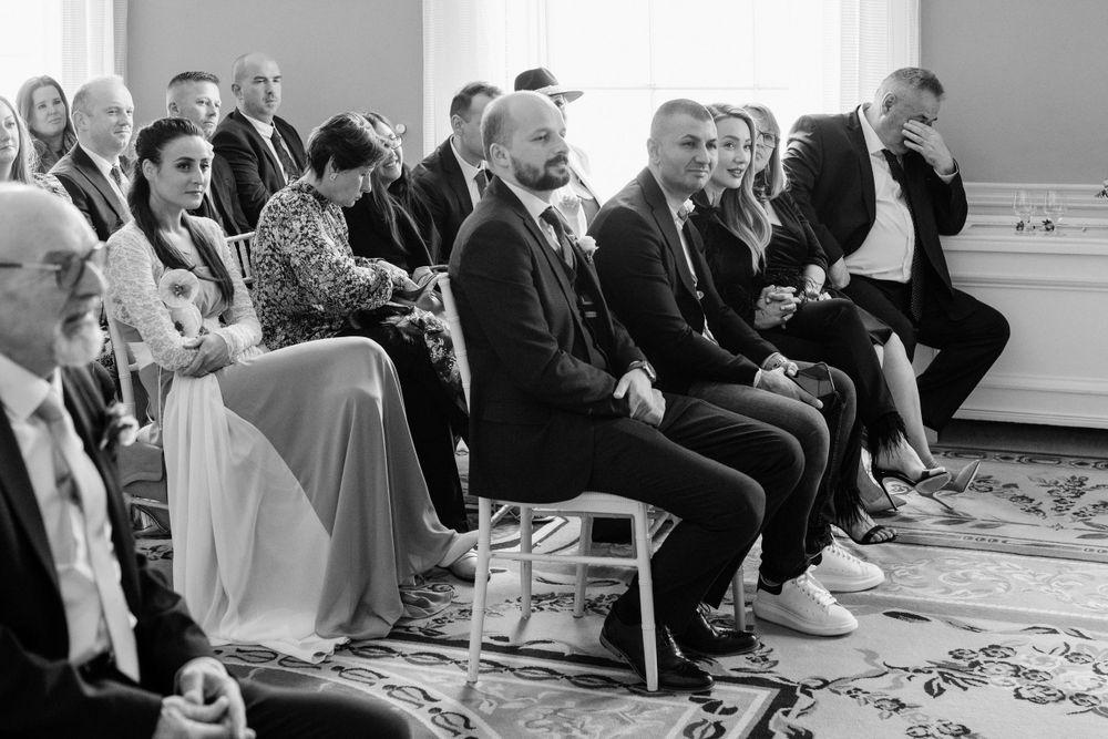 Wedding guests seated in formal attire laugh during indoor ceremony in black and white photography.