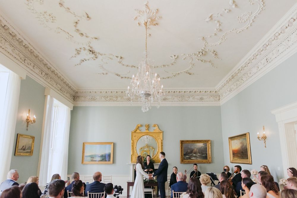 A couple exchanges vows during an elegant wedding ceremony in a historic room with ornate ceilings and mint green walls.