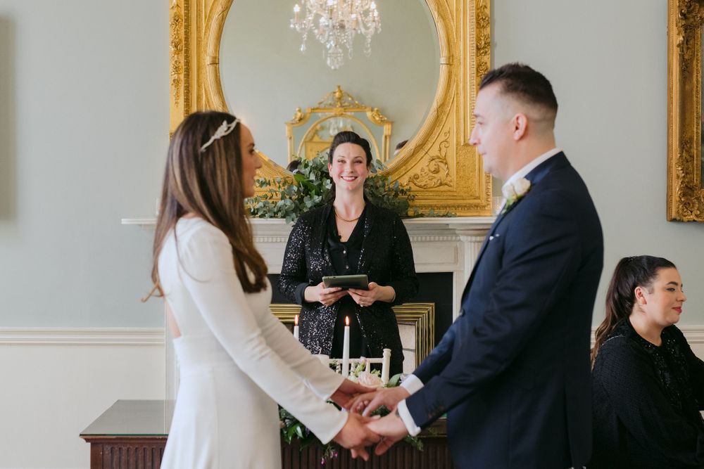 An intimate wedding ceremony takes place in an elegant room with ornate gold-framed mirrors and white walls.