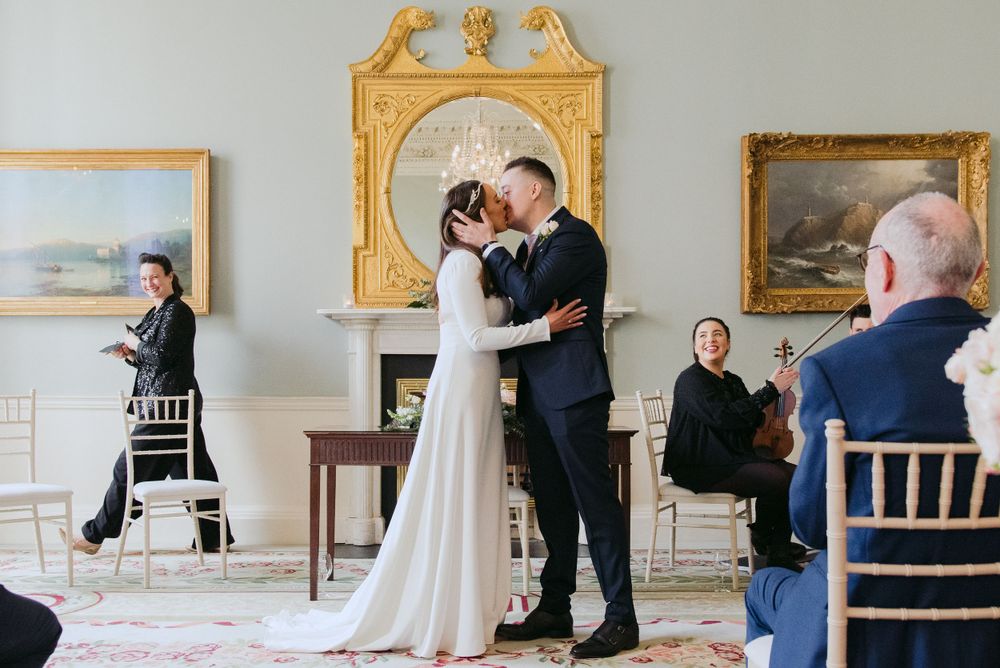 An intimate wedding ceremony takes place in an elegant room with ornate gold-framed mirrors and landscape paintings on the walls.