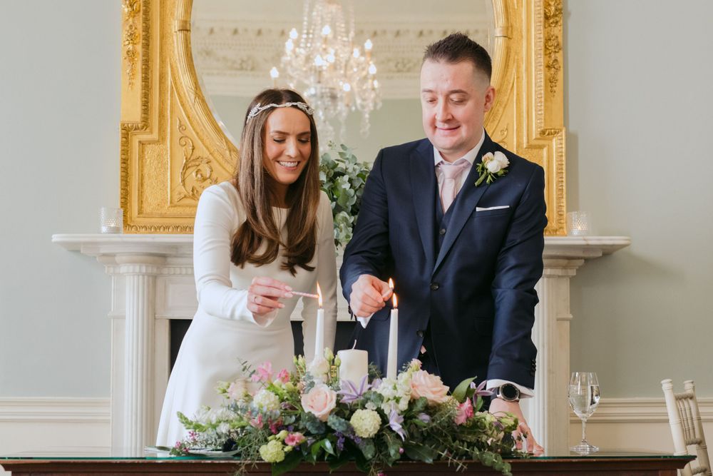 A couple lights a unity candle during their wedding ceremony in an elegant room with a gold framed mirror.