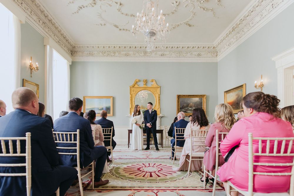 An intimate wedding ceremony takes place in an elegant white room with ornate moldings and a vintage pink carpet.