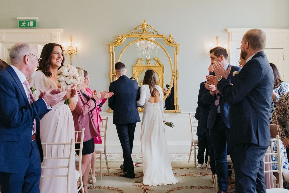 Wedding guests celebrate during an elegant ceremony in a room with ornate gold mirror and white decor.