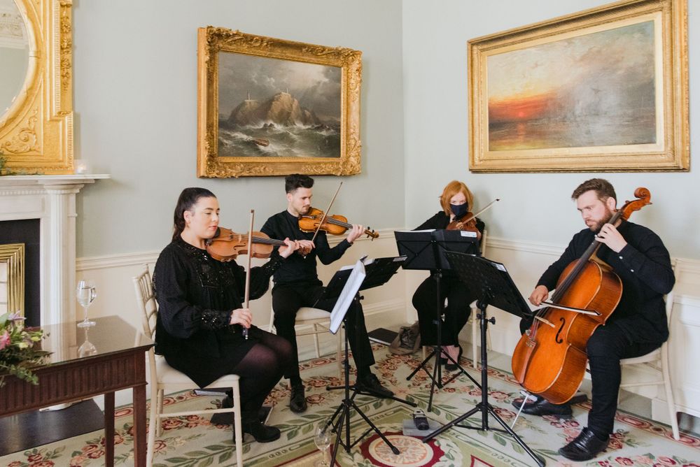 A string quartet performs classical music in an elegant room with framed paintings and a fireplace.