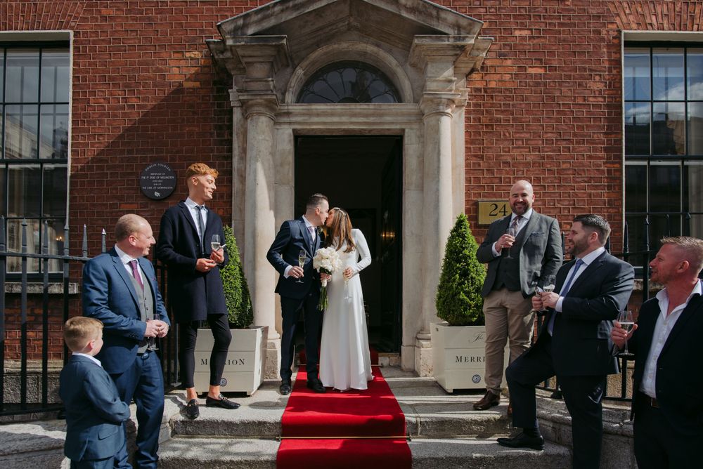 A wedding ceremony takes place on the steps of a classic red brick mansion with a formal red carpet entrance.