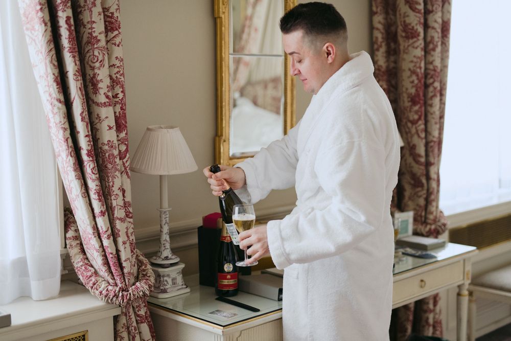Someone in a white bathrobe pours a beverage in an elegant hotel room with floral curtains and antique furniture.