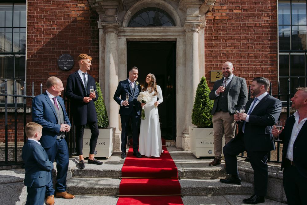 Wedding couple stands at entrance of historic brick building with red carpet and guests gathered on the steps.