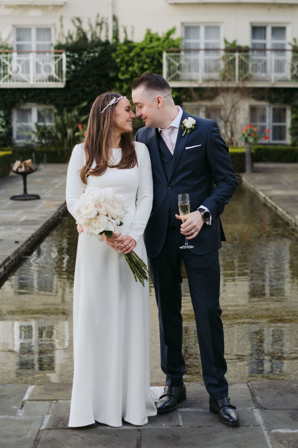 Bride in elegant white dress and groom in navy suit share intimate moment in front of reflection pool.