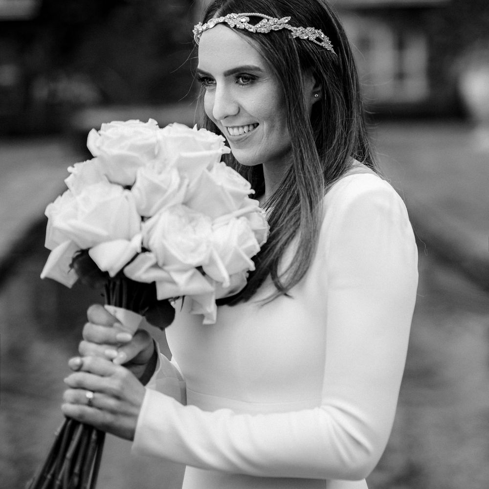 A bride in a long-sleeved white dress and delicate headpiece holds a bouquet of white roses in black and white photography.