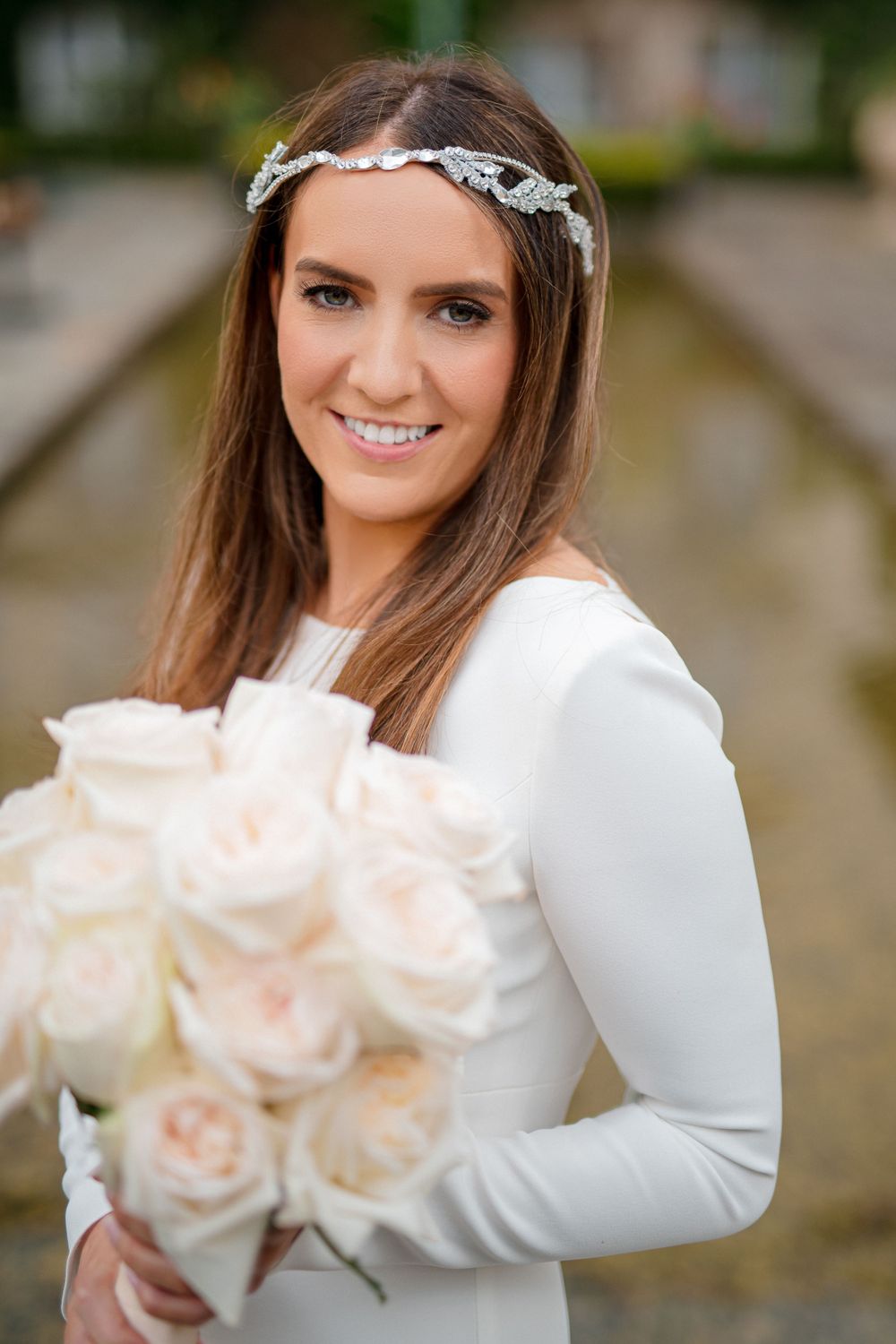 The bride holds a bouquet of pale pink roses while wearing a delicate headpiece and long-sleeved white gown.