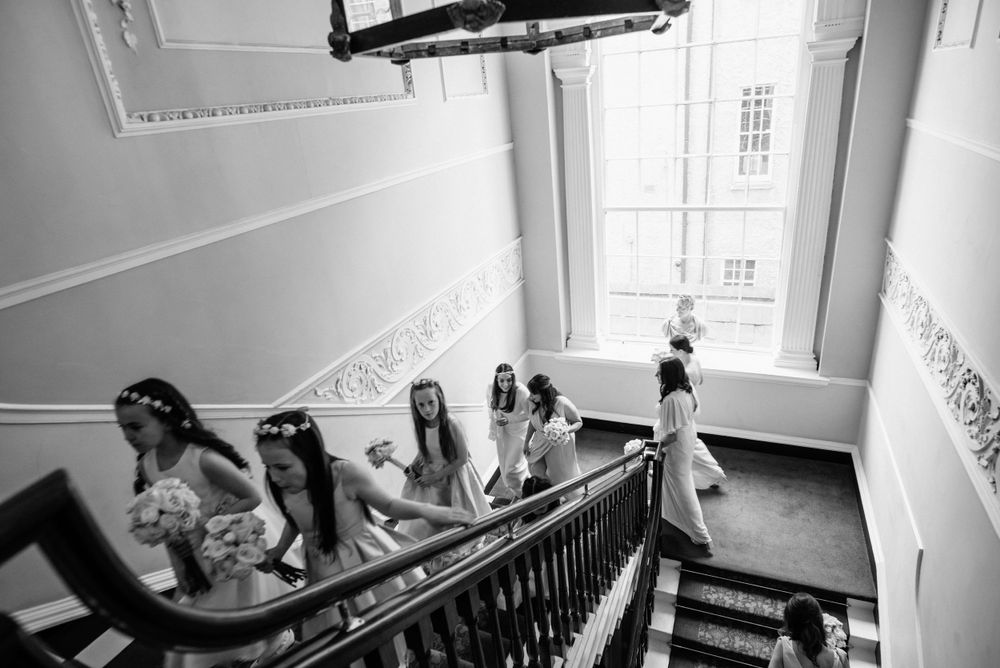 A black and white photograph shows people in formal attire descending an elegant staircase with ornate railings and moldings.