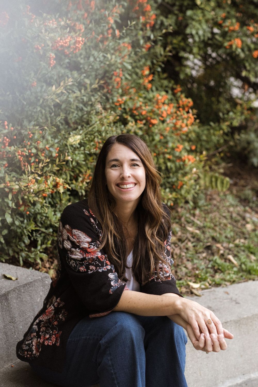 Someone wearing a black floral top and jeans sits casually on concrete steps surrounded by autumn foliage.