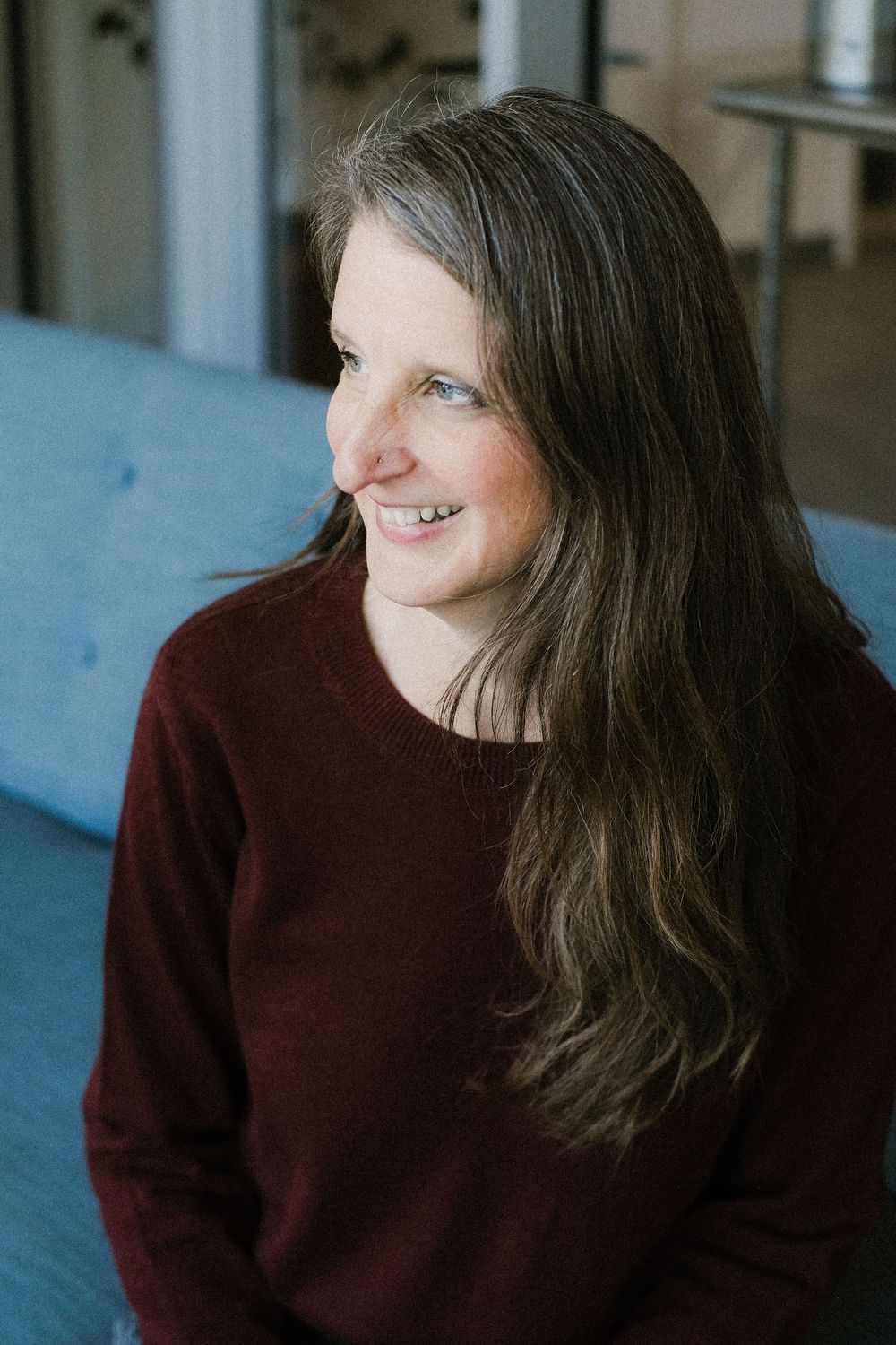 A woman with long brown hair wearing a burgundy sweater smiles while looking to the side on a blue couch.