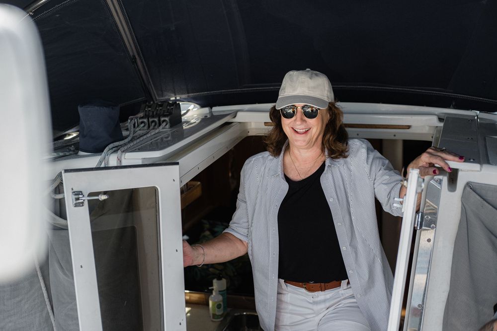 A person in a gray hat and sunglasses leans into a white boat cabin while holding onto a railing.