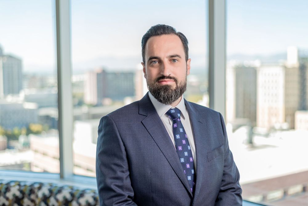Professional business person in a navy suit stands confidently by large office windows with city views.